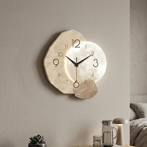 Puede incluir: Un reloj de pared con un marco irregular de textura de piedra beige y un centro circular iluminado que recuerda a la luna. La esfera presenta números arábigos negros, manecillas negras finas y un pequeño peso de péndulo circular colgante.