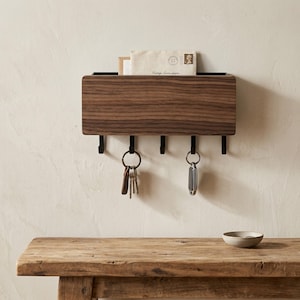 May include: A wall-mounted organiser featuring a rectangular dark wood front panel with a visible grain pattern. The unit includes a top slot for mail and five black metal hooks underneath, with two key rings hanging from the hooks.