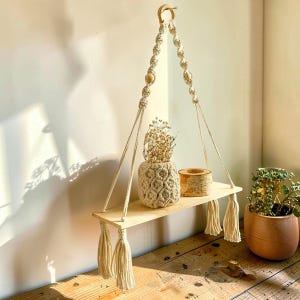 May include: A macrame hanging shelf with a light wood shelf and cream-colored tassels. The shelf is suspended by cream-colored rope and adorned with wooden beads. A small, textured vase and a wooden container sit on the shelf.