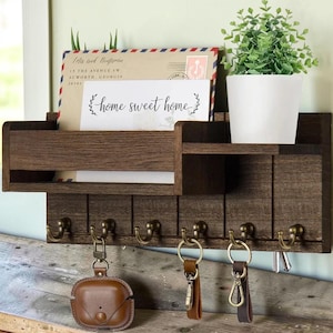 May include: A dark brown wooden wall-mounted organizer with a shelf, mail slot, and six hooks. A white pot with a green plant sits on the shelf. The mail slot holds an envelope and a card that reads "home sweet home".