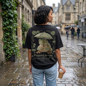 Puede incluir: Camiseta negra con un diseño de rana bajo un paraguas de lunares. La rana es de color verde claro y beige. El texto en la camiseta dice: "Prepared for rain, unprepared for small talk."