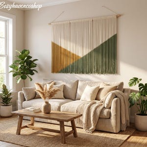 May include: A living room scene featuring a beige sofa, wooden coffee table, and a large macrame wall hanging with geometric shapes in mustard yellow and olive green. Potted plants and a woven rug complete the cozy, neutral-toned decor.