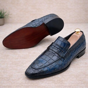 May include: A pair of blue loafers featuring a textured crocodile-embossed pattern. One shoe is positioned to show the upper with a strap across the vamp, while the other is placed to display the smooth, dark brown wooden sole and black heel.