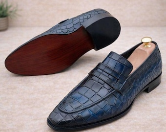 May include: A pair of blue loafers featuring a textured crocodile-embossed pattern. One shoe is positioned to show the upper with a strap across the vamp, while the other is placed to display the smooth, dark brown wooden sole and black heel.