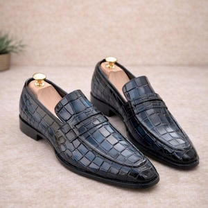 May include: A pair of dark blue loafers featuring a glossy crocodile skin texture. The shoes have a classic slip-on design with a strap across the vamp, a low heel, and a wooden shoe tree inserted into each shoe with a gold-toned metal knob.