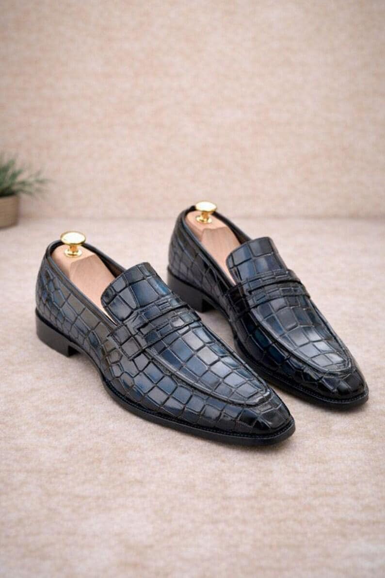 May include: A pair of dark blue loafers featuring a glossy crocodile skin texture. The shoes have a classic slip-on design with a strap across the vamp, a low heel, and a wooden shoe tree inserted into each shoe with a gold-toned metal knob.