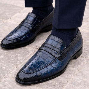 May include: A pair of navy blue loafers featuring a glossy crocodile-embossed texture, a classic penny strap across the vamp, and a low heel. The shoes are worn with dark navy socks and dark trousers, showcasing a polished finish and structured design.