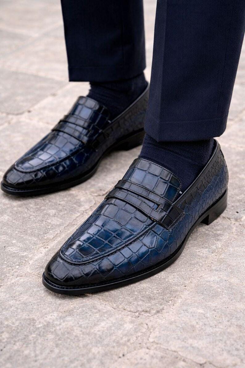 May include: A pair of navy blue loafers featuring a glossy crocodile-embossed texture, a classic penny strap across the vamp, and a low heel. The shoes are worn with dark navy socks and dark trousers, showcasing a polished finish and structured design.