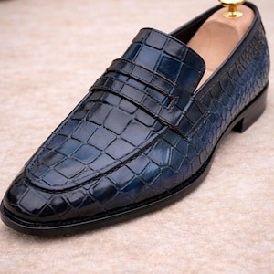 May include: A navy blue leather loafer featuring a glossy crocodile skin texture. The shoe has a classic penny strap across the vamp, a rounded toe, and a black sole. A wooden shoe tree with a gold-toned knob is inserted into the heel area.