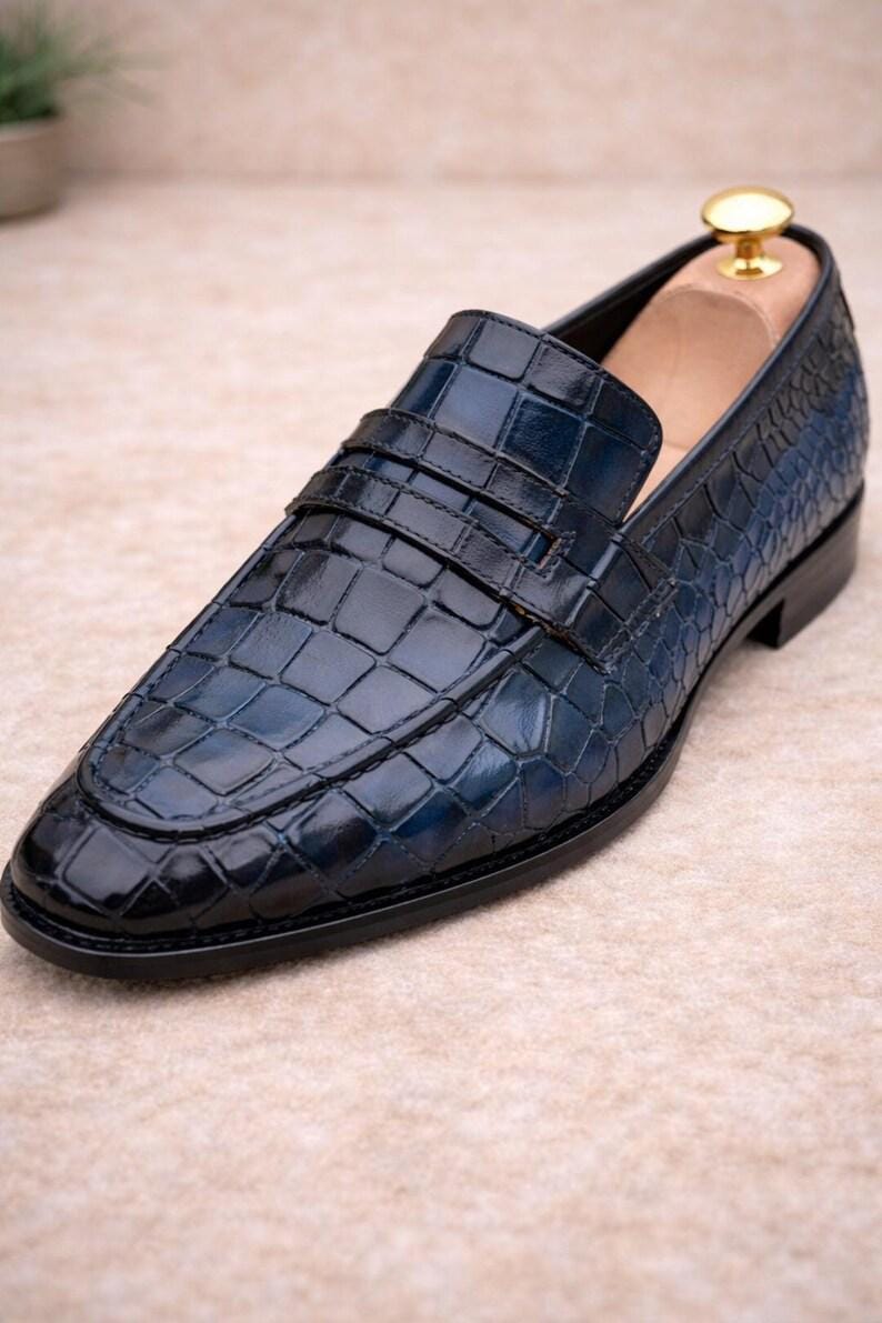 May include: A navy blue leather loafer featuring a glossy crocodile skin texture. The shoe has a classic penny strap across the vamp, a rounded toe, and a black sole. A wooden shoe tree with a gold-toned knob is inserted into the heel area.