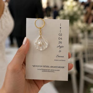 Op de afbeelding: Een heldere, schelpvormige sleutelhanger met een parel in het midden hangt aan een gouden ring. De sleutelhanger is bevestigd aan een witte kaart met tekst en data. De kaart luidt "SEVGI EN GUZEL ANAHTARDIR."