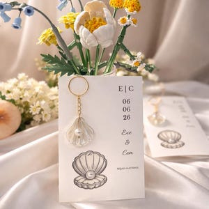 Puede incluir: Un recuerdo de boda decorativo con un llavero en forma de concha con un acento de perla. La tarjeta incluye las iniciales "E|C" y la fecha "06 06 26", junto con los nombres "Ece & Cem" y una ilustración de concha. Un pequeño ramo de flores está en el fondo.