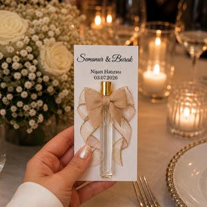 Puede incluir: Un vial de perfume de vidrio transparente con una tapa dorada está adherido a una tarjeta blanca con los nombres "Semanur & Burak" y la fecha 03.07.2026. La tarjeta está decorada con un lazo beige y una cinta dorada. El fondo incluye flores, velas y cristalería.