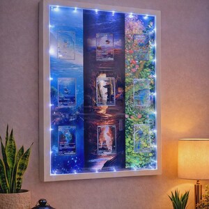Può includere: Un espositore con cornice bianca con nove carte collezionabili, illuminato da piccole luci bianche. Le carte presentano varie scene d'arte fantasy. Il display è montato su una parete viola chiaro, con una pianta in vaso e una lampada in primo piano.