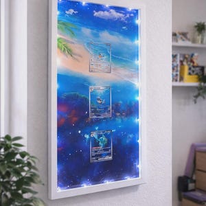 Può includere: Opera d'arte con cornice bianca con tre carte collezionabili Pokémon su uno sfondo vibrante di oceano e cielo. La cornice è illuminata da piccole luci bianche. L'opera è montata su una parete bianca.