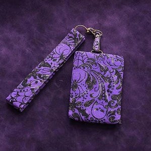 May include: A purple key fob set with a matching wristlet. The set features a decorative black and silver pattern with spiderwebs and swirls. The key fob has a metal clasp and the wristlet is a long, rectangular shape.