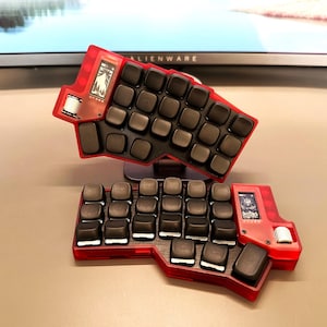 Peut inclure: Un clavier ergonomique divisé avec un cadre rouge translucide et des touches noires. Chaque moitié possède un petit écran et une molette blanche. Le clavier est conçu pour le confort et l'efficacité, avec une disposition unique.