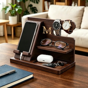 Può includere: Un organizer da scrivania in legno marrone scuro con più scomparti. È dotato di un supporto per telefono, porta orologi, un ripiano per occhiali e un vassoio per piccoli oggetti. Un telefono è in carica nel supporto. L'organizer è personalizzato con il nome "MICHAEL".