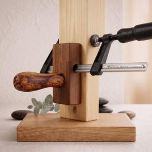 May include: A light-colored wooden vertical post mounted on a rectangular wooden base, featuring a dark brown wooden block with a polished burl wood handle and a metal Stahlwerk clamp attached to the side, securing the components together.
