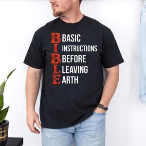 Könnte beinhalten: Schwarzes T-Shirt mit dem Wort "BIBLE" in Rot, vertikal gestapelt, mit den Worten "BASIC INSTRUCTIONS BEFORE LEAVING EARTH" in Weiß. Das T-Shirt wird von einer Person getragen.