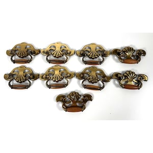 Könnte beinhalten: Eine Sammlung von neun antiken Schubladengriffen aus Messing. Jeder Griff hat ein verziertes, stilisiertes Design mit einem Holzgriff. Das Messing hat eine warme, gealterte Patina. Die Griffe sind auf einer weißen Oberfläche angeordnet.