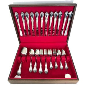 May include: A wooden silverware chest with a red velvet interior, showcasing a complete set of polished silver flatware. The set includes knives, forks, spoons, and serving utensils, all neatly arranged. The chest is open, revealing the contents.