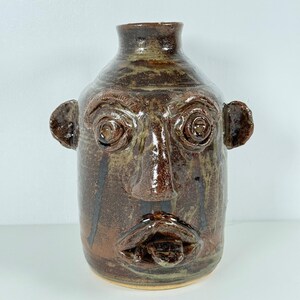 May include: A brown ceramic face jug with a detailed face, including eyes, a nose, and a mouth. The jug has two ear-like handles and a narrow neck. The pottery has a rustic, handmade appearance.