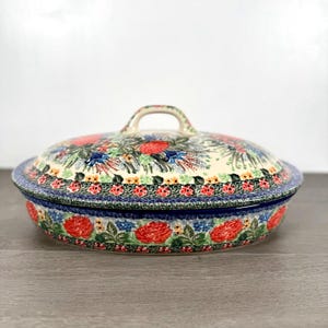 May include: A round ceramic casserole dish with a matching lid, featuring a cream-colored base decorated with vibrant red floral patterns, green leaves, and small blue accents. The rim and lid handle are detailed with a dark blue decorative border.