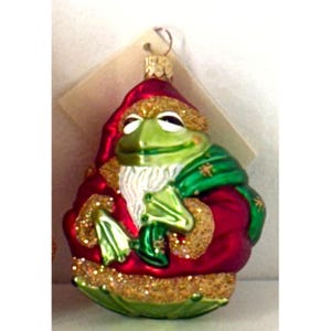 May include: A Christmas ornament shaped like a green frog wearing a red Santa suit with gold glitter accents. The frog has a white beard and is holding a green sack. The ornament has a gold metal loop for hanging.