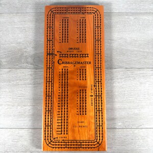 May include: A rectangular wooden cribbage board with a natural light brown finish. The board features black printed markings, including the text Drueke Model 1950, Cribbagemaster, and Game 121 Points, along with rows of holes for scoring.