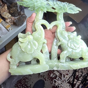 May include: A light green carved stone sculpture featuring two elephants standing on a base, each positioned beneath a palm tree. The elephants have their trunks raised and curved toward each other, forming a heart shape between the two trees.