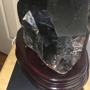 May include: A large, dark, faceted crystal point resting on a polished, oval, dark brown wooden base. The crystal and base are positioned on a silver Accuteck digital scale, which displays a weight measurement of 9.105 pounds on its screen.