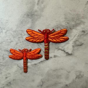 May include: Two embroidered dragonfly patches in orange and gold. The larger dragonfly is about 2 inches long and the smaller dragonfly is about 1 inch long.