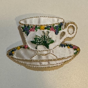 May include: A white teacup and saucer with a gold trim and floral embroidery. The embroidery features pink, yellow, blue, and green flowers.