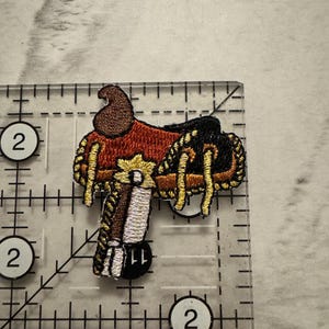 May include: Embroidered patch of a Western saddle, featuring brown, red, black, gold, and white thread. The patch is approximately 2 inches in length and width, with intricate stitching details. The design is a miniature representation of a classic saddle.