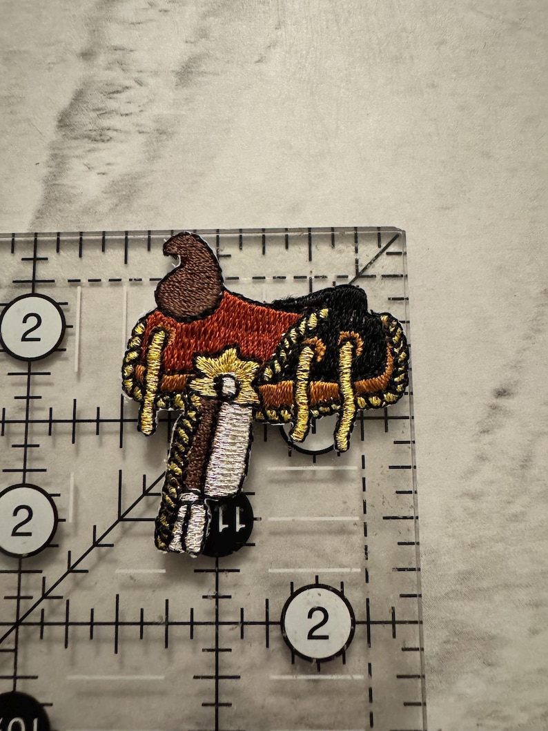 May include: Embroidered patch of a Western saddle, featuring brown, red, black, gold, and white thread. The patch is approximately 2 inches in length and width, with intricate stitching details. The design is a miniature representation of a classic saddle.