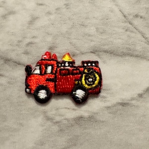 May include: Embroidered patch of a red fire truck with black and white wheels. The patch features a yellow bell and a black outline. The patch is designed for application on clothing or accessories.
