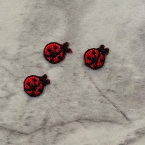 May include: Three small, embroidered ladybug patches. Each patch features a red body with black spots and a black head. The patches are arranged on a light gray, textured surface.