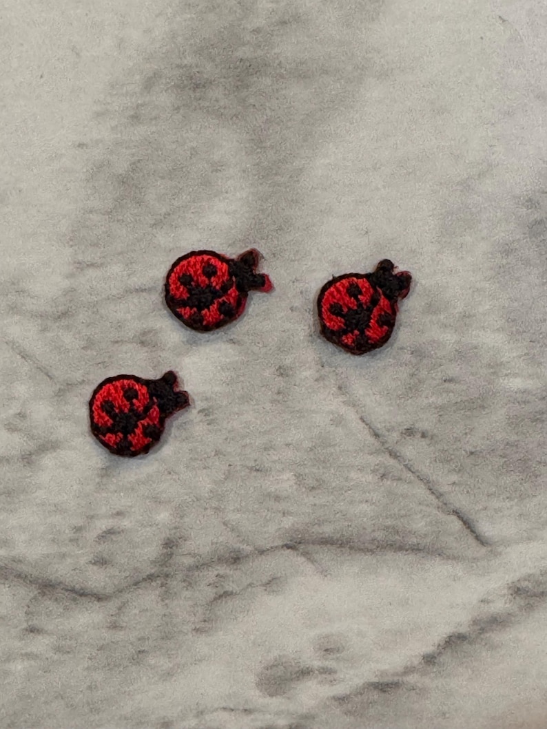 May include: Three small, embroidered ladybug patches. Each patch features a red body with black spots and a black head. The patches are arranged on a light grey, textured surface.