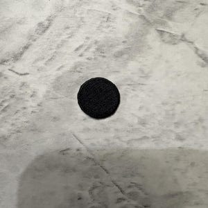 May include: A small, circular black patch with a textured, embroidered surface. The patch features a clean, stitched edge and a uniform, solid black color throughout, presenting a simple and functional design for various textile applications.