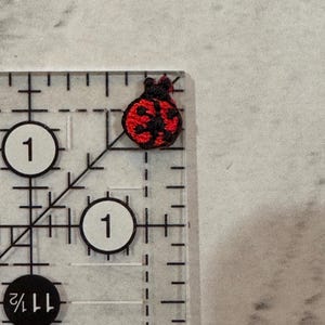 May include: A miniature embroidered ladybird patch, featuring a red body with black spots and head. The patch is placed on a clear ruler, displaying measurements in inches. The ladybird is a symbol of good fortune, often used in crafting projects.