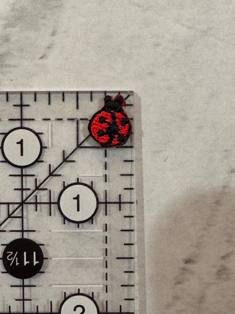 May include: A miniature embroidered ladybird patch, featuring a red body with black spots and head. The patch is placed on a clear ruler, displaying measurements in inches. The ladybird is a symbol of good fortune, often used in crafting projects.