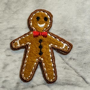 May include: A felt gingerbread man applique with a brown outline and a red bow tie. The gingerbread man has a smiling face with black eyes and a white stitched mouth. The applique is designed for crafts and holiday decorations.