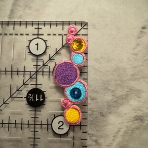 K&ouml;nnte beinhalten: Gestickter Aufn&auml;her mit einer Reihe farbenfroher, kreisf&ouml;rmiger Designs. Der Aufn&auml;her enth&auml;lt einen gro&szlig;en lila Kreis, einen blauen Kreis mit einer zentralen Perle und kleinere gelbe und rosa Kreise. Der Aufn&auml;her ist etwa 5 cm lang.