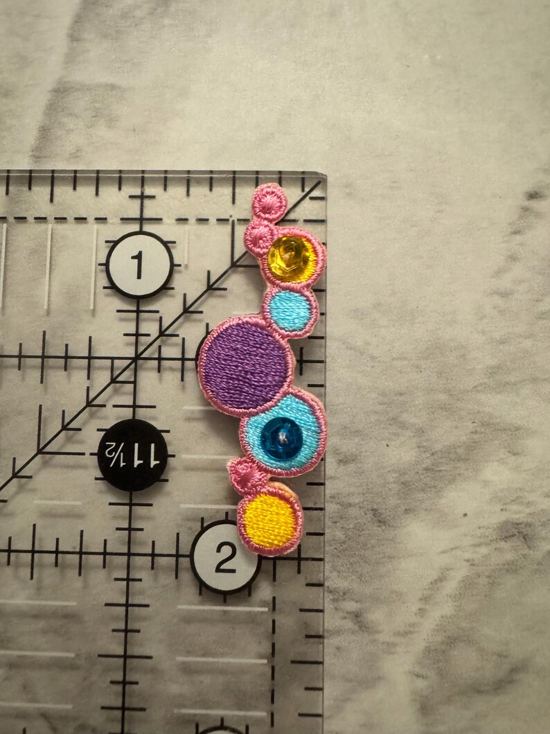 K&ouml;nnte beinhalten: Gestickter Aufn&auml;her mit einer Reihe farbenfroher, kreisf&ouml;rmiger Designs. Der Aufn&auml;her enth&auml;lt einen gro&szlig;en lila Kreis, einen blauen Kreis mit einer zentralen Perle und kleinere gelbe und rosa Kreise. Der Aufn&auml;her ist etwa 5 cm lang.