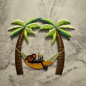 May include: Embroidered patch featuring a monkey relaxing in a hammock strung between two palm trees. The monkey is black and brown, the hammock is yellow, and the palm trees are brown and green. The patch is designed for clothing or accessories.