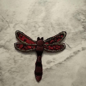 May include: A red and black dragonfly-shaped applique. The wings are a deep red with black accents and gold trim. The body is a textured, striped pattern in shades of red and black. This decorative applique can be used for crafts and sewing projects.