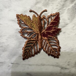 May include: An embroidered butterfly patch featuring a central body and wings composed of intricate leaf-like structures. The embroidery uses a color palette of metallic copper, deep burgundy, olive green, and golden yellow threads with openwork details.