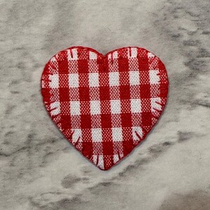 May include: A heart-shaped patch with a red and white gingham pattern. The patch has a stitched edge and is set against a gray and white marbled background. This decorative patch is suitable for crafts and sewing projects.