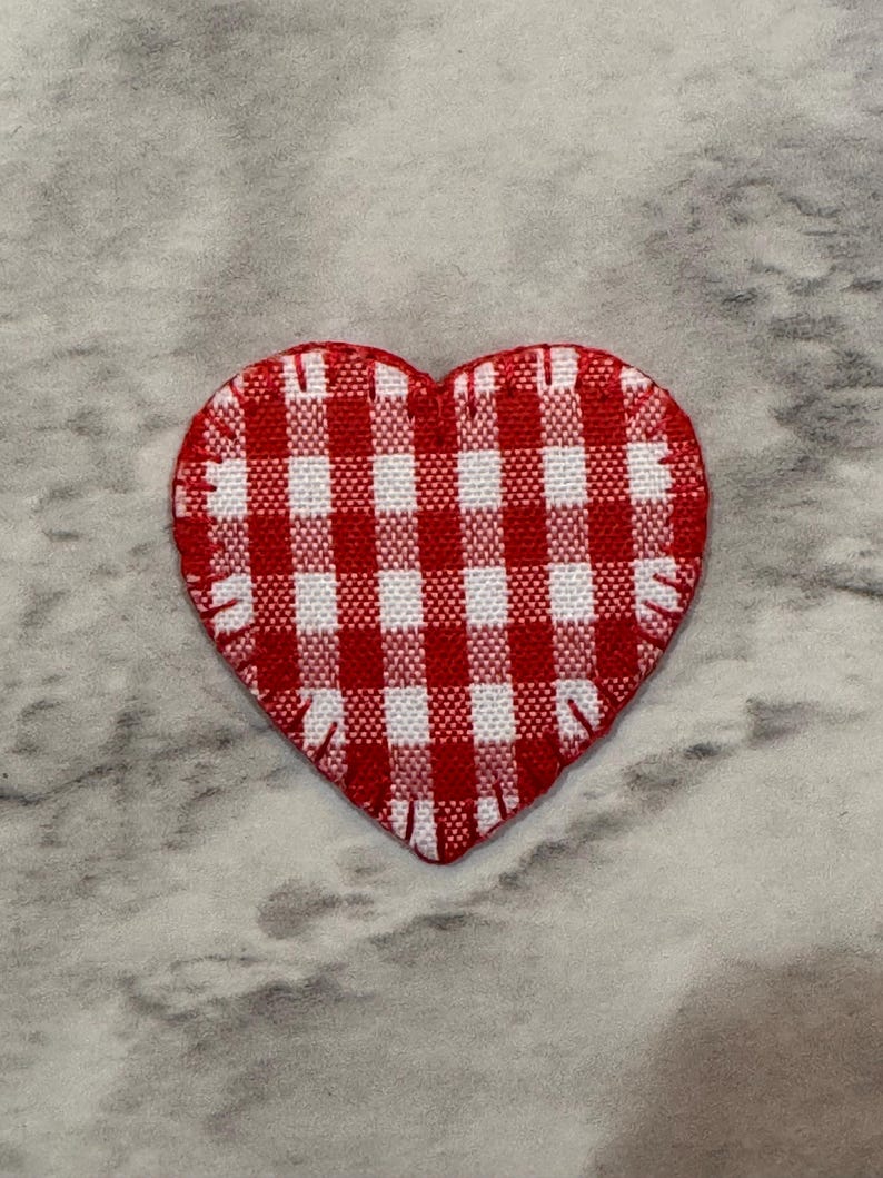 May include: A heart-shaped patch with a red and white gingham pattern. The patch has a stitched edge and is set against a gray and white marbled background. This decorative patch is suitable for crafts and sewing projects.